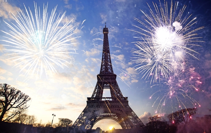 Ein Bild zeigt den Eiffelturm bei Sonnenuntergang umgeben von buntem Feuerwerk am Himmel.
