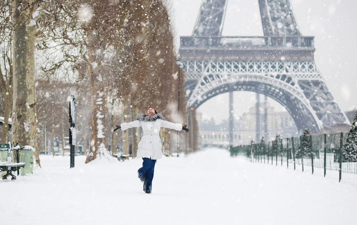 Eine Person steht mit ausgebreiteten Armen im Schnee vor dem Eiffelturm in Paris im Winter.