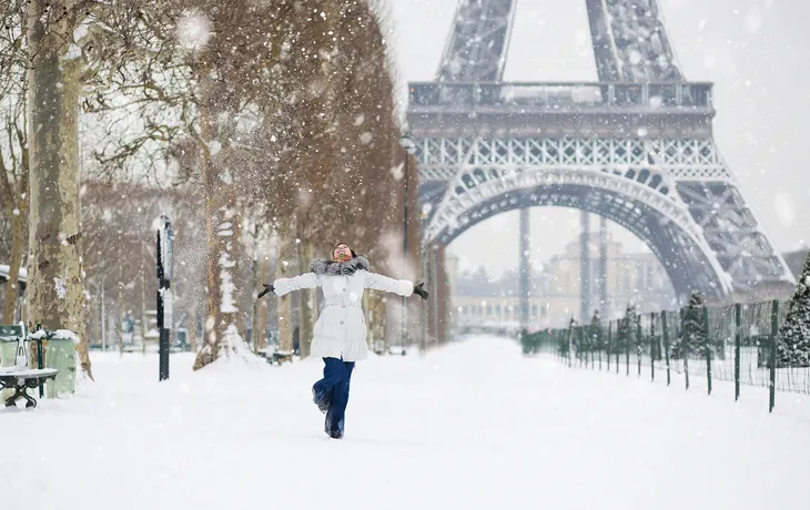 Eine Person steht mit ausgebreiteten Armen im Schnee vor dem Eiffelturm in Paris im Winter.
